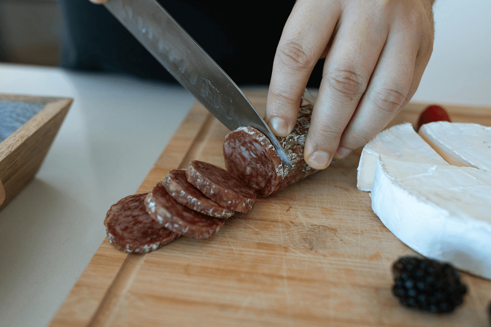 Sliced artisan salami on a wooden cutting board with brie cheese and berries, being prepared for a charcuterie-style snack.